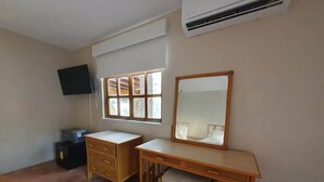 Ein Zimmer mit Schreibtisch, Kommode, Fernseher und einem Fenster mit Jalousien.