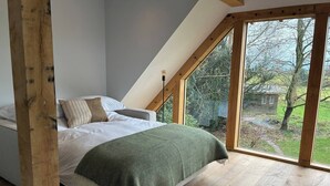 A bedroom with a sloped ceiling, a bed with white linens, a green throw blanket, and a view of a garden through large windows. - The Loft – 1 bed space with gated parking (Binfield Heath)