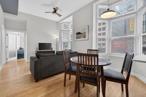 Een moderne woonkamer met een grijze bank, een houten eettafel en stoelen, een televisie en grote ramen met zonwering.