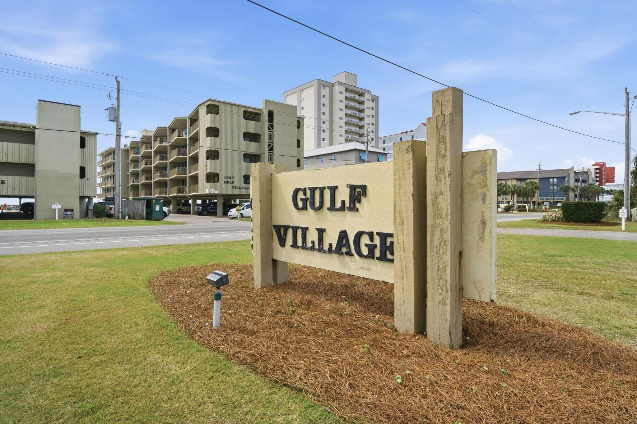 Letrero que indica 'Gulf Village', con edificios de varios pisos y una carretera de fondo.