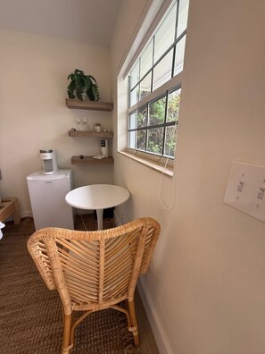 Quarto pequeno com uma mesa branca, cadeira de vime, frigorífico branco, estantes com objetos e uma janela com vista.