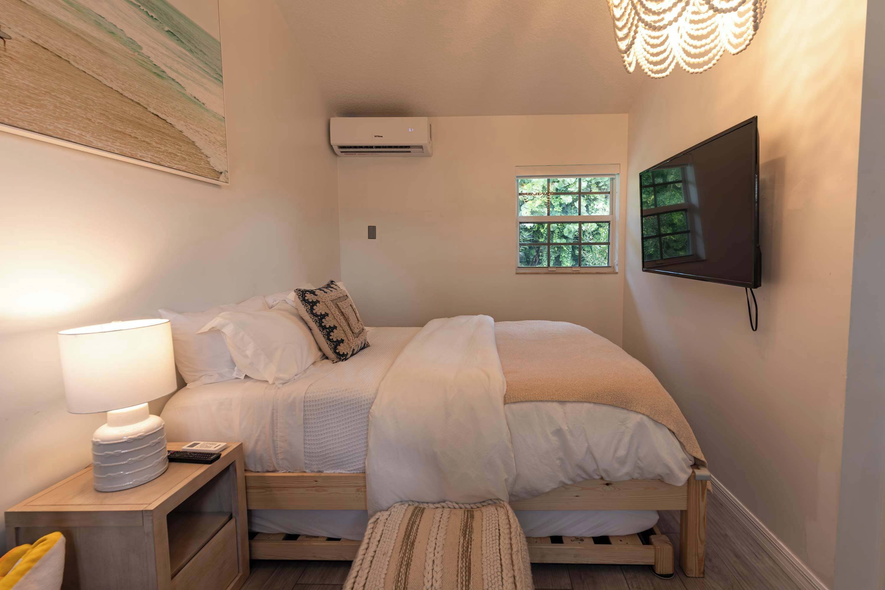 A bedroom with a bed, bedside lamp, TV, air conditioning unit, window, and a wall-mounted light fixture.
