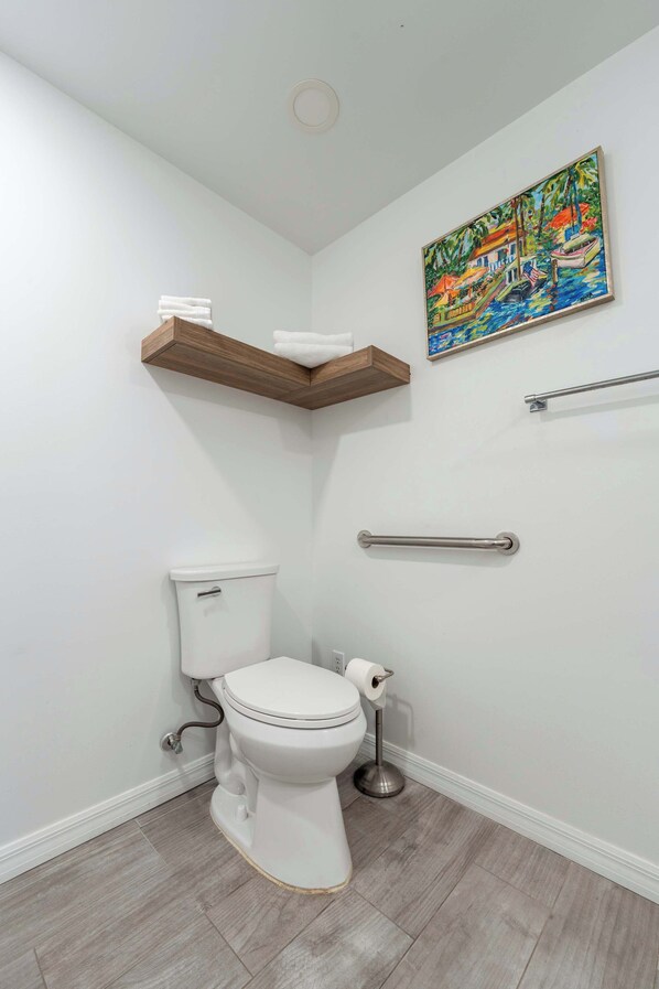 A bathroom with a toilet, a wooden shelf, a handrail, and a painting on the wall.