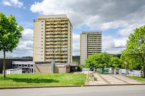 A multi-story building with a flat roof, a parking area with a few vehicles, and a grassy area with trees. - Cozy Holiday Apartment in the Harz with Shared Pool and Balcony (Braunlage)