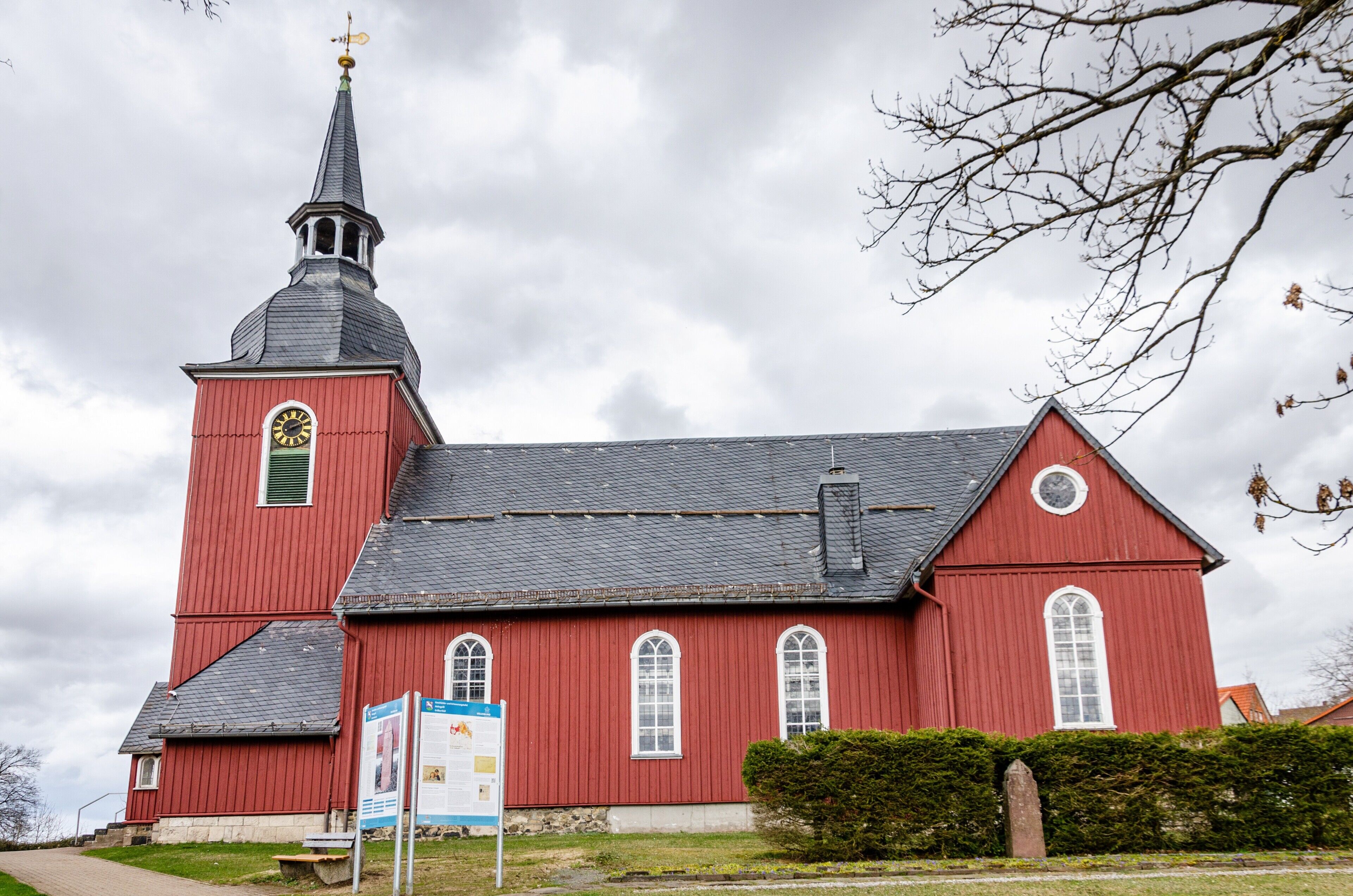 Eine rote Kirche mit einem Kirchturm und einer Uhr, ein Weg und ein Hinweisschild.