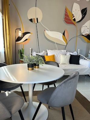 Ein modernes Wohnzimmer mit einem runden weißen Tisch, grauen Stühlen, einer weißen Couch mit Kissen und einer Wand mit abstrakten Blattmustern.