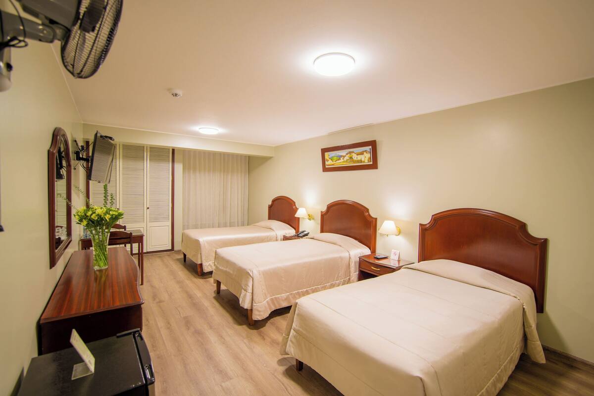 A hotel room with three beds, a desk, a mirror, and a picture on the wall.