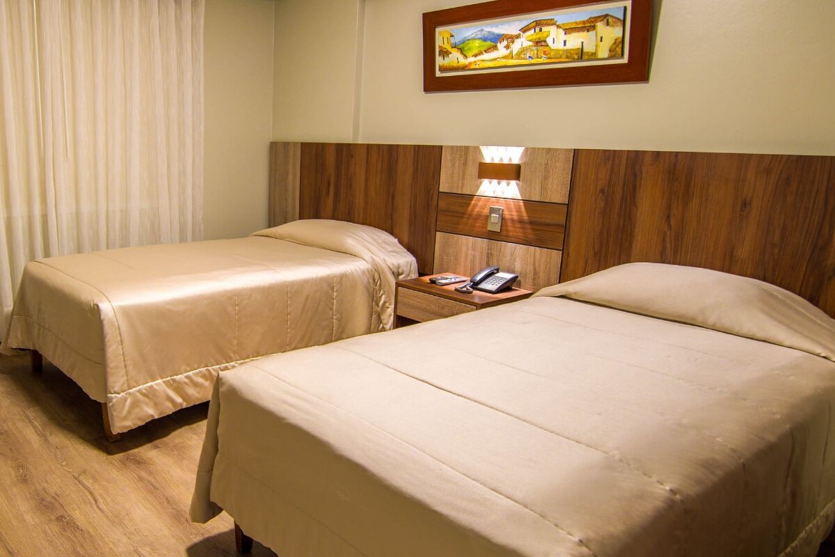 A hotel room with two beds, a wooden desk, a chair, a vase with flowers, and a painting on the wall.
