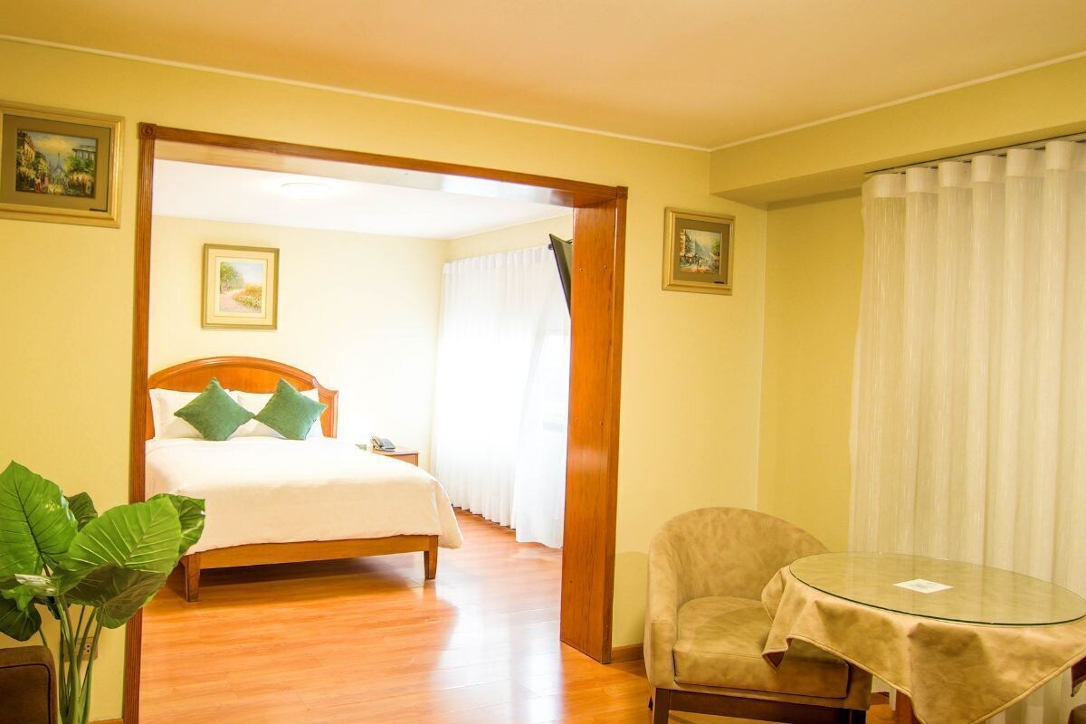 A hotel room with a bed, a chair, a small table, and a plant.