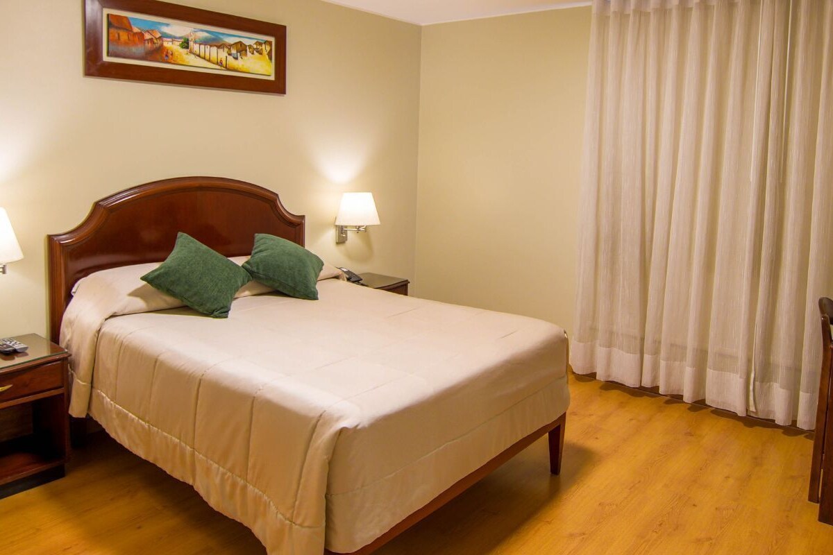A hotel room with a bed, bedside lamps, a desk, a chair, and a painting on the wall.