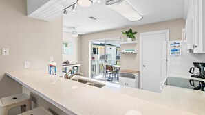A modern kitchen with a white countertop, stainless steel sink, and a refrigerator. There is a dining area with a table and chairs visible through a glass door.