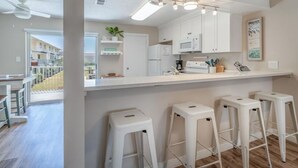 A modern kitchen with white cabinets, a microwave, and a coffee machine. There are four white bar stools and a ceiling fan.