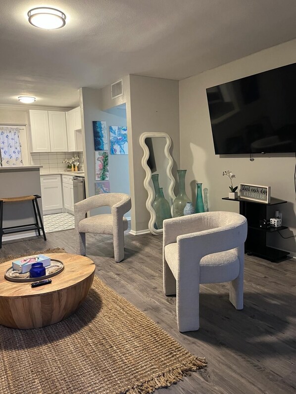 A living room with two armchairs, a coffee table, a flat-screen TV, and a kitchen in the background.