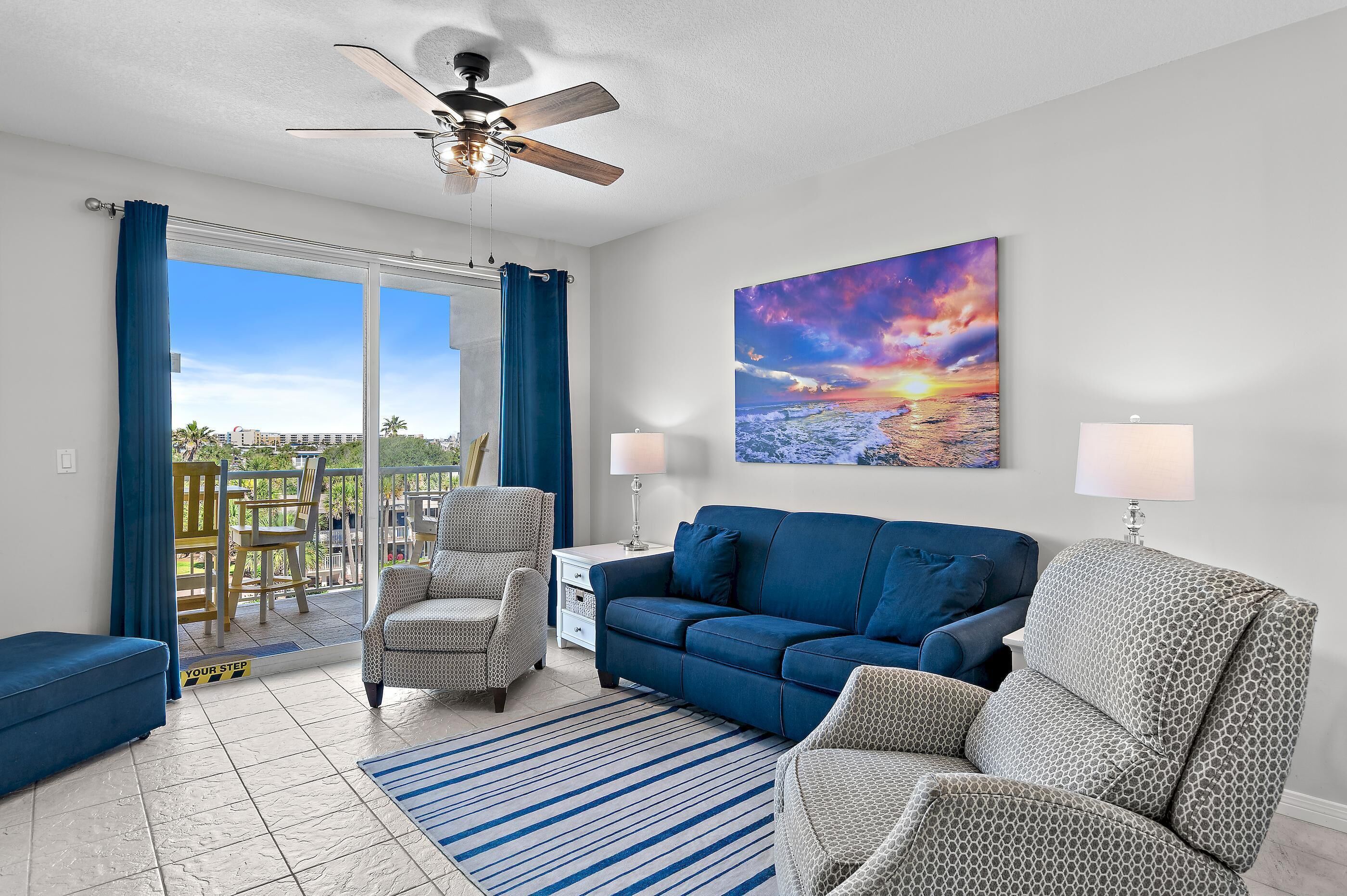 A living room with a sofa, chairs, a ceiling fan, a painting, and a view through a glass door.