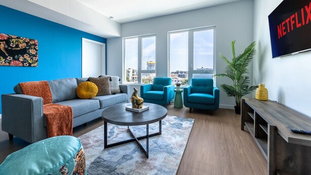 Ein modernes Wohnzimmer mit einem grauen Sofa, blauen Sesseln, einem Couchtisch und einem großen Fenster mit Blick auf die Stadt.