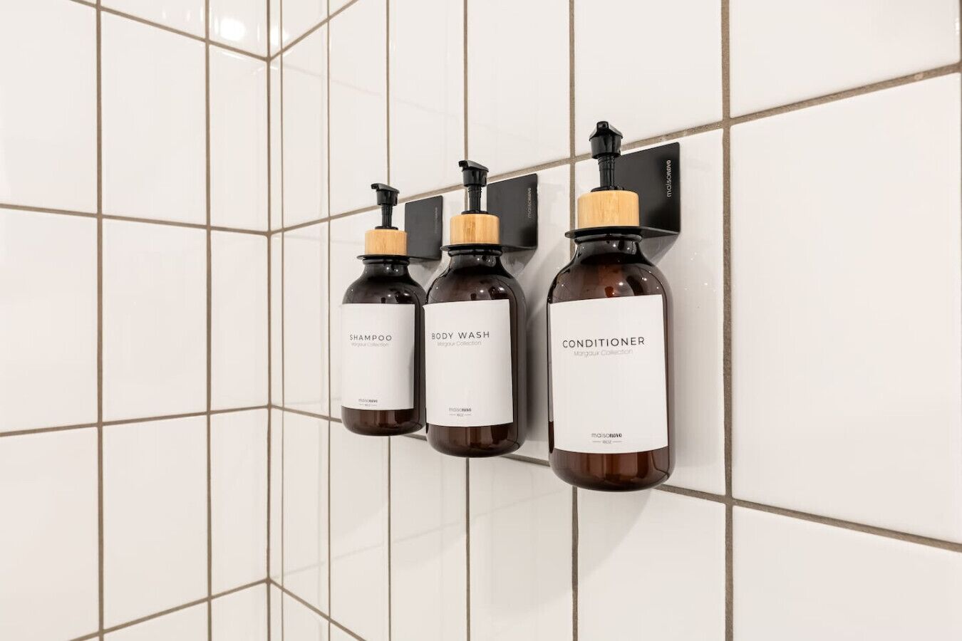 Three bottles of shampoo, body wash, and conditioner mounted on a tiled wall.