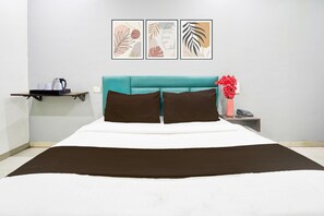 A hotel room with a bed, a teal headboard, a nightstand with a phone and a vase of flowers, and a shelf with cups and a kettle. - Super Hotel O Dindoli Surat (Surat)