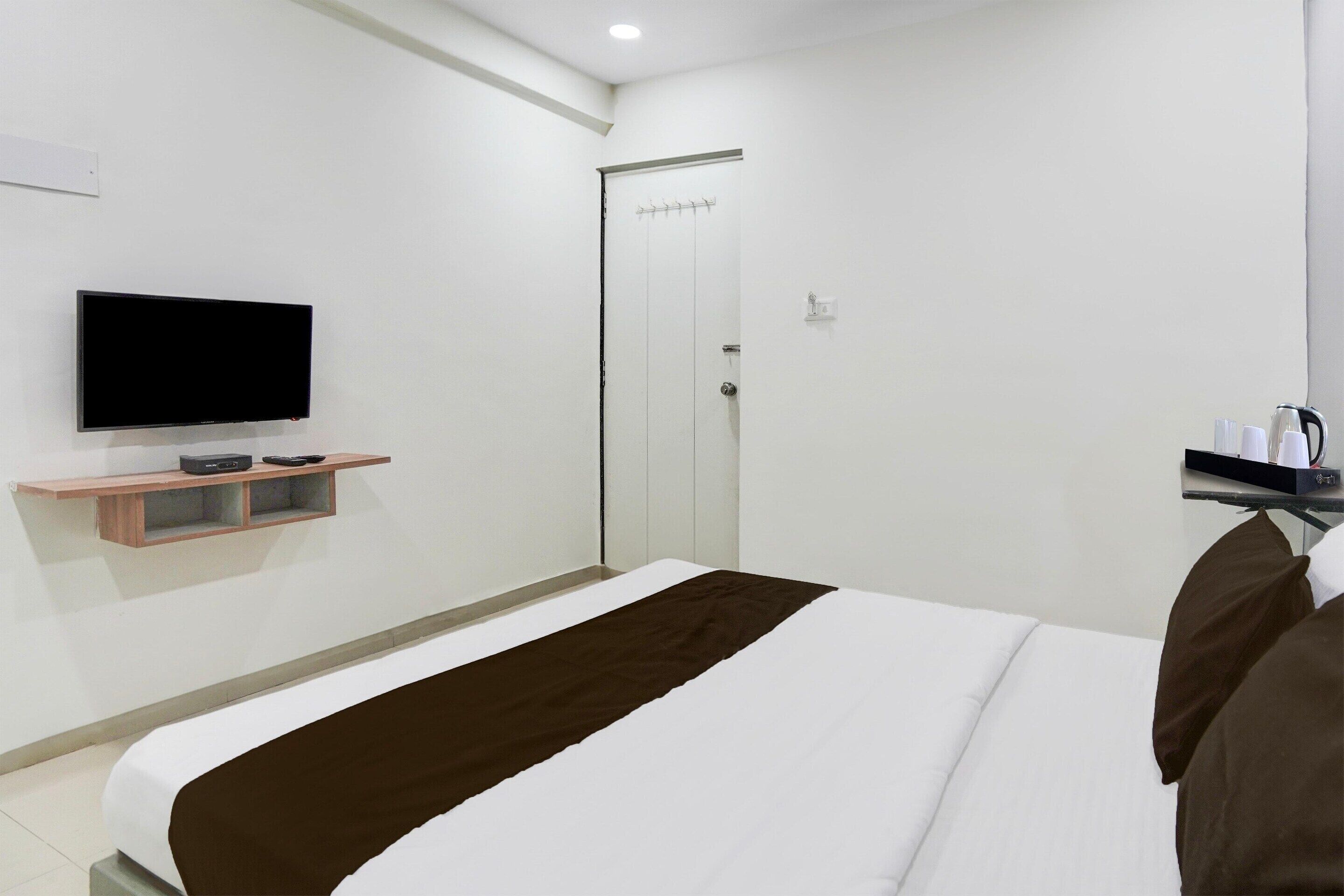 Habitación de hotel con una cama, un televisor montado en la pared, una puerta y un estante pequeño con objetos encima.
