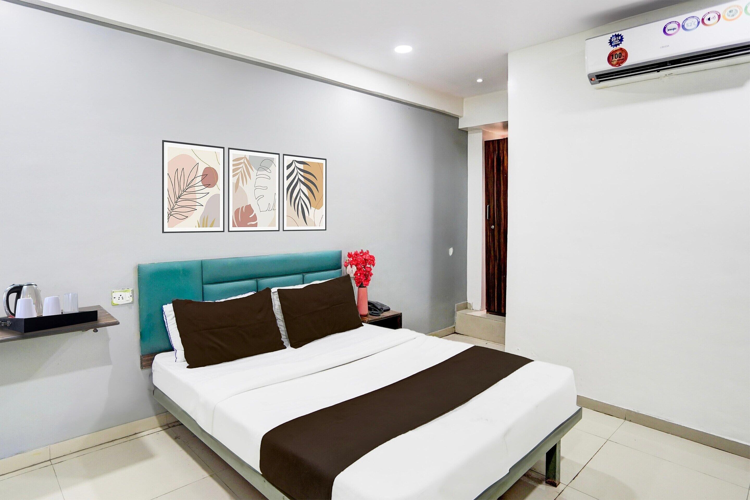 A neatly made bed with a teal headboard, a bedside table with a vase of red flowers, and three wall-mounted abstract art pieces.