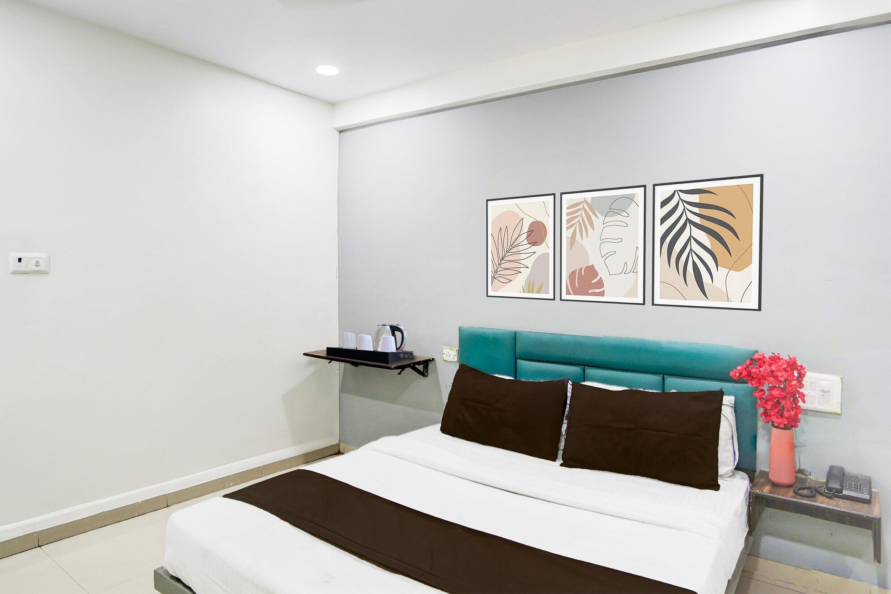Une chambre d’hôtel bien rangée, avec un lit, une table de chevet, une étagère avec des tasses et une bouilloire, et trois œuvres d’art encadrées accrochées au mur.
