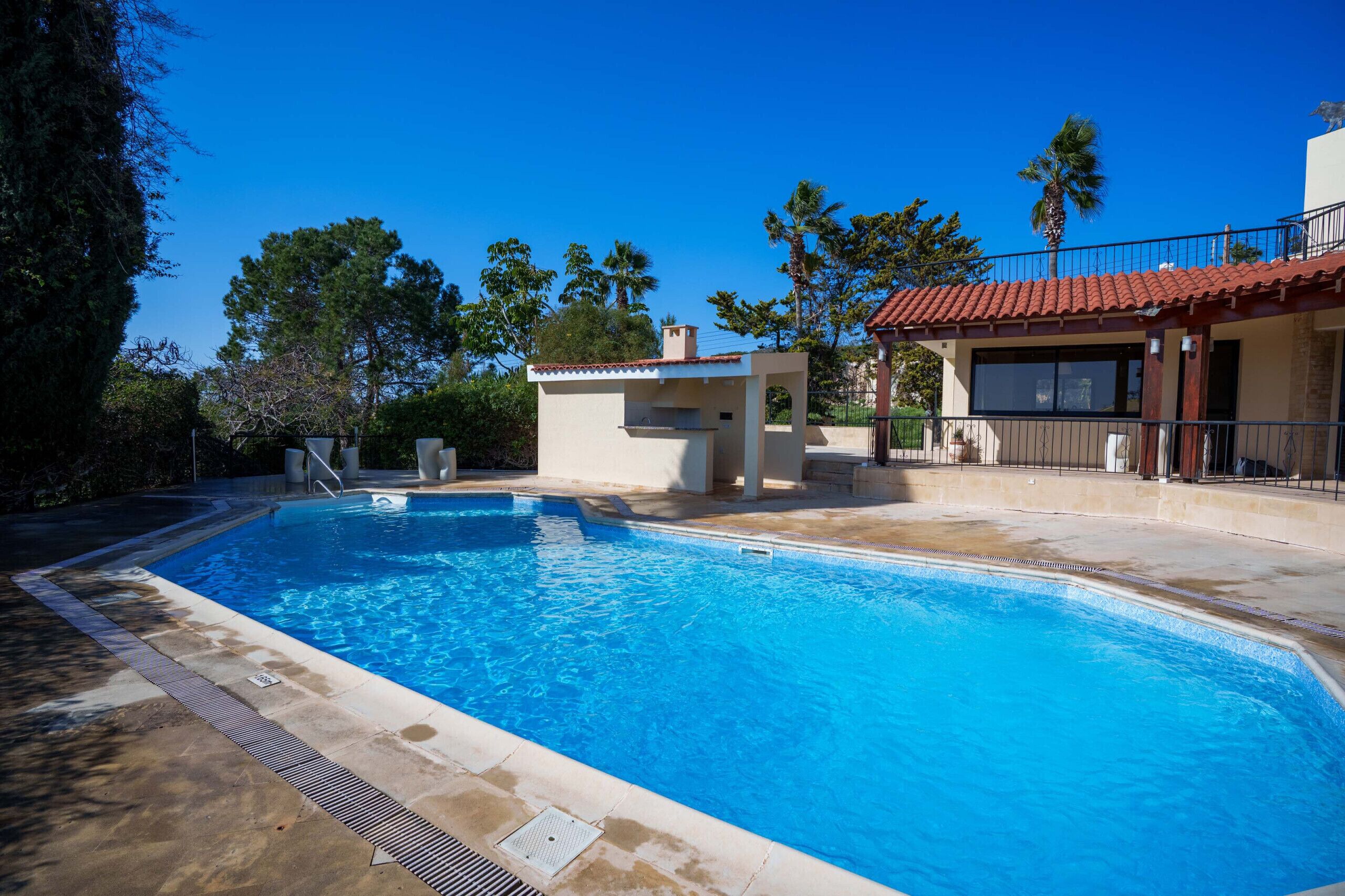 Une piscine avec un petit bâtiment attenant, entourée d’arbres et d’un autre bâtiment dont le toit est recouvert de tuiles rouges.