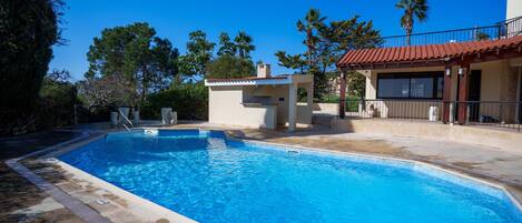 Une piscine avec un petit bâtiment attenant, entourée d’arbres et d’un autre bâtiment dont le toit est recouvert de tuiles rouges.