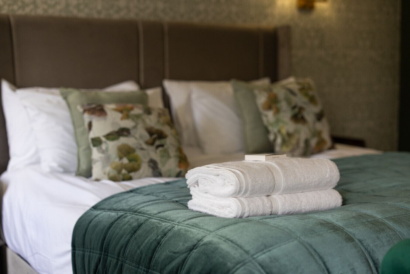 Una cama bien hecha con un edredón verde, almohadas blancas y una toalla doblada.