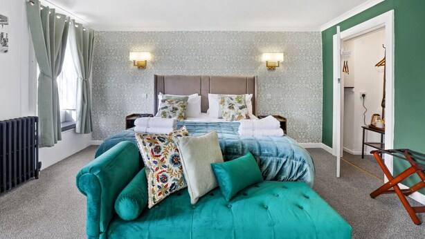 A bedroom with a bed, two wall-mounted lights, a radiator, a window with curtains, and a green wall. - The Drawing Room at The Merchants (Berwick-Upon-Tweed)