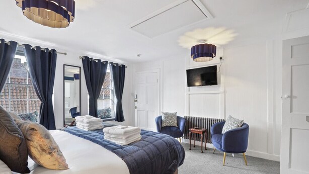 A bedroom with a bed, two blue armchairs, a radiator, a TV, and a window with curtains. - The Morning Room at The Merchants (Berwick-Upon-Tweed)