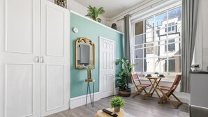 Een kamer met een muur in turquoise, witte deuren, een spiegel, een klein tafeltje met een afstandsbediening, en een eethoek met stoelen en een tafel.