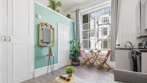 Een moderne woonkamer met een teal accentmuur, een eethoek met tafel en stoelen, een keuken met vaatwasser, en een groot raam met uitzicht op gebouwen.
