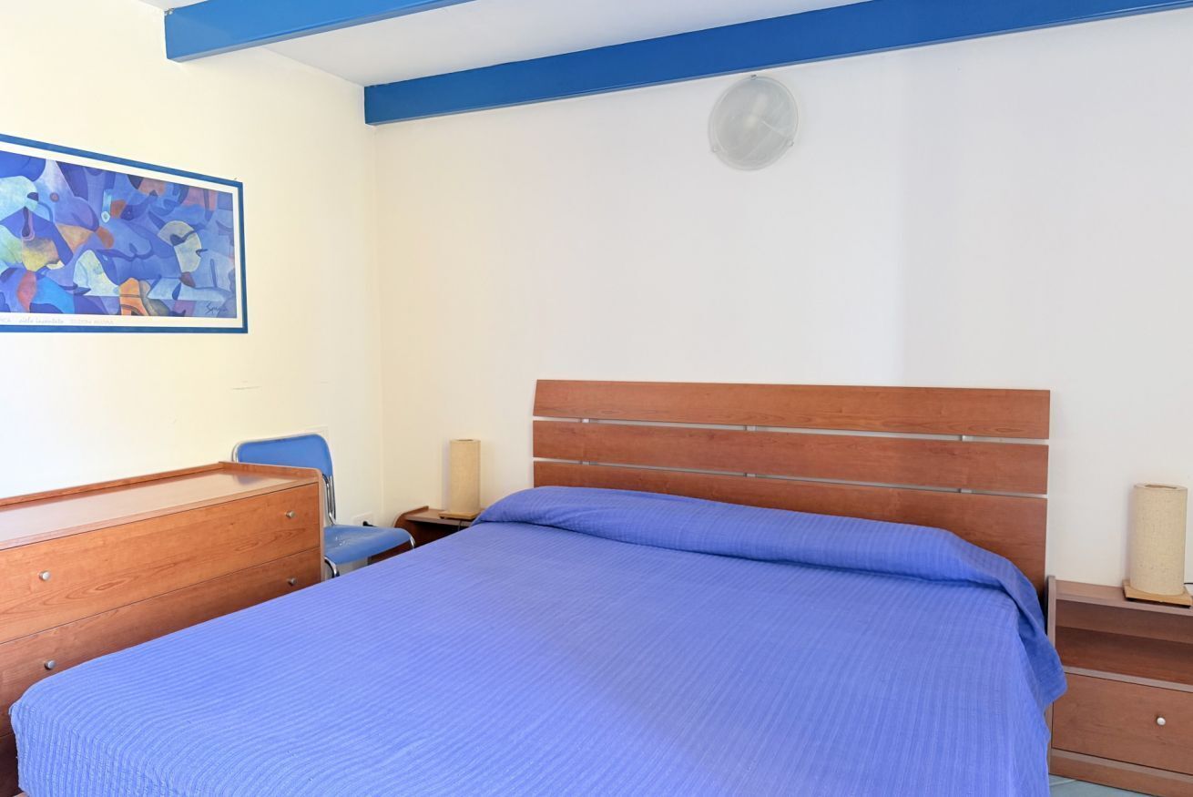 A bedroom with a wooden headboard, a blue bedspread, a wooden dresser, a chair, and a wall clock.