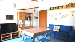 A room with a wooden table, blue chairs, a blue sofa, a bookshelf, a television, and wall decorations including fish and star patterns. - S361 – Sirolo, apartment in a residence with swimming pool (Sirolo)