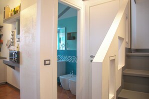 Una cocina con refrigerador, microondas y repisas con objetos. Se ve un baño con inodoro y lavamanos a través de una puerta abierta.