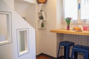 A corner of a room with a white wall, a wooden shelf with books and a cactus, and two blue stools. - Casa Zeda WelcHome (Intra)