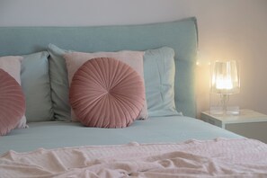 Una cama con cabecera color turquesa, varios almohadones, una lámpara de noche y una manta rosa.