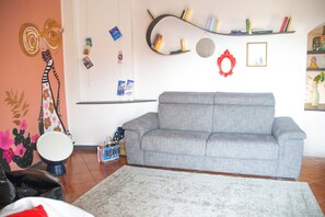 A living room with a grey sofa, a patterned rug, a guitar, and a wall with a cactus mural and shelves with books and decorative items. - Casa Zeda WelcHome (Intra)