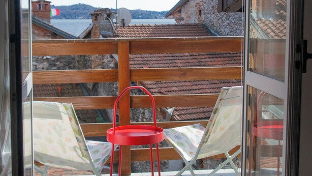A balcony with a view of a body of water and buildings, a wooden railing, and two red chairs. - Casa Zeda WelcHome (Intra)