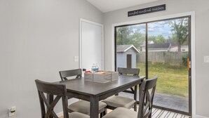 Un coin repas avec une table et des chaises, une porte-fenêtre coulissante donnant sur un jardin, et un panneau accroché au mur.