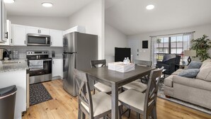 Une cuisine moderne équipée d’appareils électroménagers en acier inoxydable, un coin repas avec une table et des chaises en bois, et un salon avec un canapé et une plante.