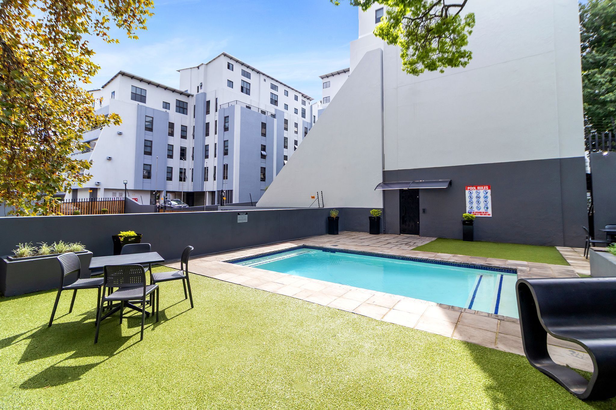 Una piscina con zona de patio, una mesa con sillas y vista a edificios modernos al fondo.