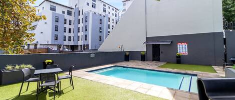 Una piscina con zona de patio, una mesa con sillas y vista a edificios modernos al fondo.