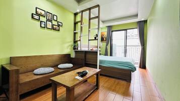 A room with a wooden bench, a small table, a bed, a shelf with items, and a window with curtains.