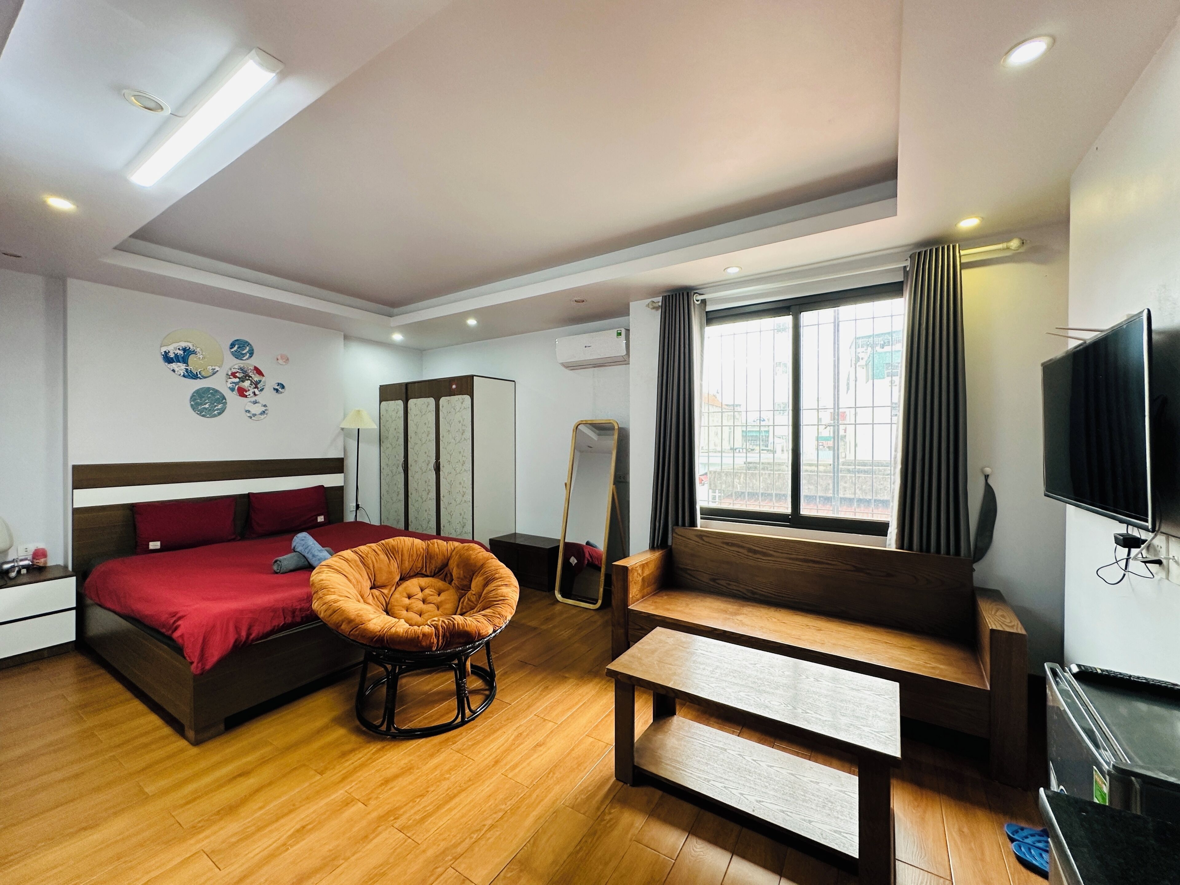 A modern hotel room with a bed, a wooden bench, a chair, a TV, a mirror, and a window with curtains.