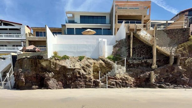 Een gebouw aan de kust met een trap die naar een balkon leidt, een zandstrand op de voorgrond en andere gebouwen op de achtergrond.