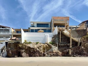 Een gebouw aan de kust met een trap die naar een balkon leidt, een zandstrand op de voorgrond en andere gebouwen op de achtergrond.
