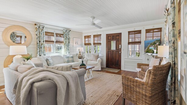 A spacious living room with a ceiling fan, large windows with bamboo blinds, a round mirror, and a wicker chair. - Two Palms Cottage on St Simons (St Simons Island)