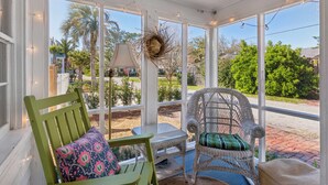 A covered porch with wicker furniture, a glass table, and a view of a garden and street. - Two Palms Cottage on St Simons (St Simons Island)