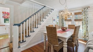 A dining area with a white table, wicker chairs, a chandelier, and a staircase with white railings. - Two Palms Cottage on St Simons (St Simons Island)