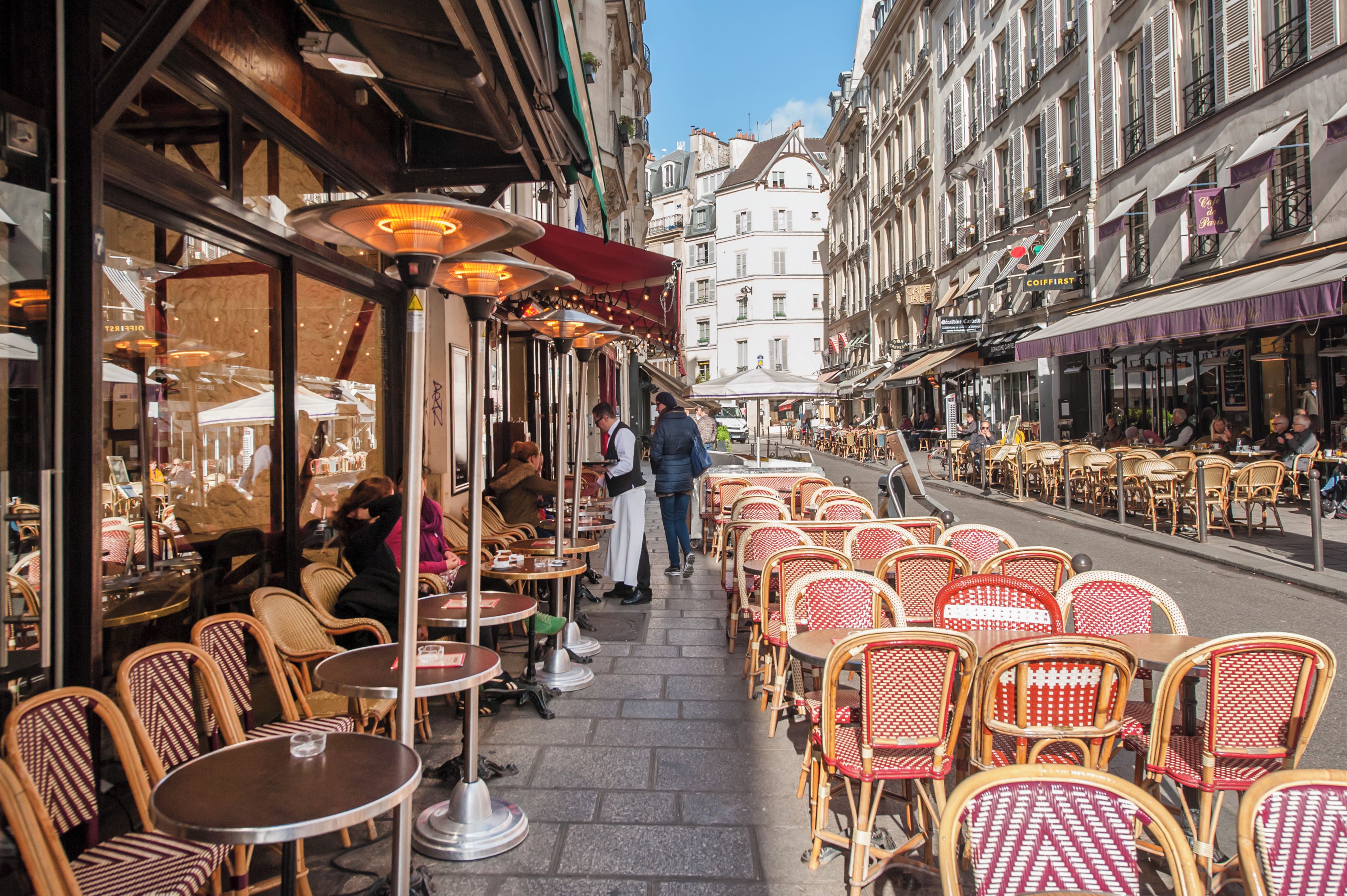 Outdoor cafe seating with tables and chairs, a street view with buildings, and a clear sky.